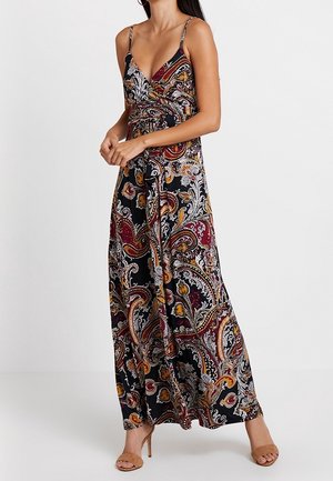 Femme portant une robe maxi sans manches à motifs cachemire multicolores avec fines bretelles et sandales à talons hauts couleur nude, debout devant un fond uni.