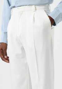 White trousers made of a lightweight fabric with pleats, featuring side pockets. Paired with a light blue button-up shirt.