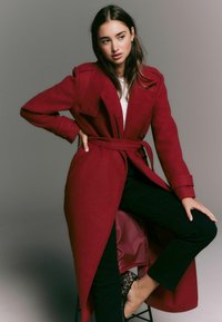 Woman wearing a long red belted coat, black pants, and leopard print shoes, seated with one leg raised against a plain gray background.