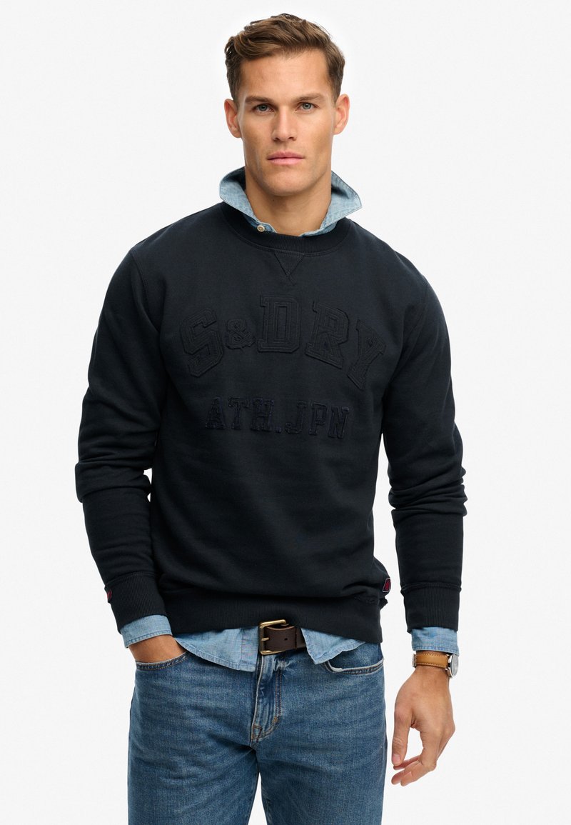 Mørkeblå sweatshirt med tekstureret logo, rund halsudskæring og ribbede manchetter, lagdelt over en lyseblå denimskjorte, der matcher med blå jeans.
