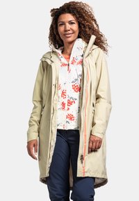 Beige wasserdichte Jacke mit Kapuze, ausgestattet mit einem korallenfarbenen Reißverschluss und einem floral gemusterten weißen Innenfutter. Getragen über einer navyfarbenen Hose, zeigt einen lässigen Schnitt.