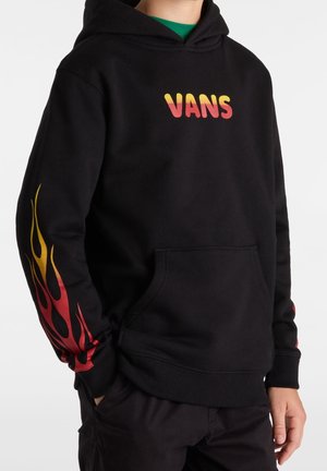 Zwarte hoodie met een gele naar rode verloopkleurige "VANS"-logo op de borst en een bijpassend vlammenontwerp op de mouwen, gedragen met de handen in de zakken.