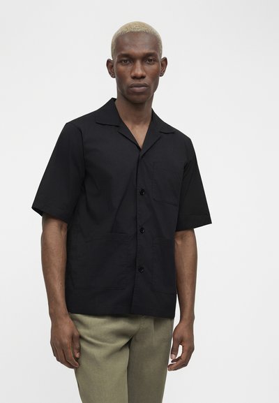 Άνδρας με κοντά ξανθά μαλλιά, ντυμένος με μαύρο πουκάμισο με κοντό μανίκι και χακί παντελόνι, στέκεται μπροστά σε απλό λευκό φόντο.