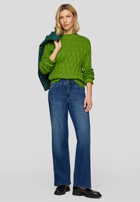 Grüner Strickpullover mit Zopfmuster und langen Ärmeln, kombiniert mit blauen, weit geschnittenen Jeans und schwarzen Loafers. Eine karierte Jacke ist über die Schulter gelegt.