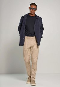 Navy jacket, black shirt, light beige pants, and tan shoes. Model wears oversized dark sunglasses, standing with hands in pockets.