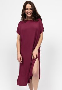 Uśmiechnięta kobieta z długimi brązowymi włosami, ubrana w luźną bordową sukienkę z wysokim rozcięciem, stojąca na tle jasnego, jednolitego tła.