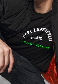 Černá prošívaná bunda s oranžovým podšívkou přes černé tričko s bílým a neonově zeleným textem. Text obsahuje "KARL LAGERFELD PARIS."