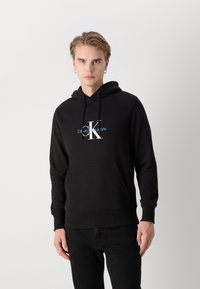 Felpa nera con cappuccio e tasca frontale. Presenta il logo bianco "CK" con il testo blu "Calvin Klein Jeans" sottostante. Tessuto dalla consistenza liscia.