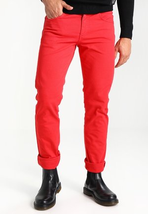 Homme portant un pantalon slim rouge vif retroussé aux chevilles, associé à des bottines en cuir noir et un haut à manches longues noir.