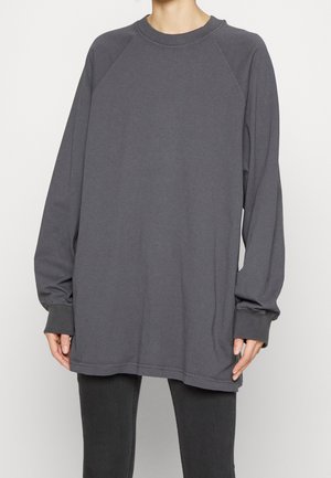 Osoba ubrana w za dużą, ciemnoszarą bluzę z długim rękawem i czarne spodnie, stojąca z rękami swobodnie opuszczonymi wzdłuż ciała na białym tle.