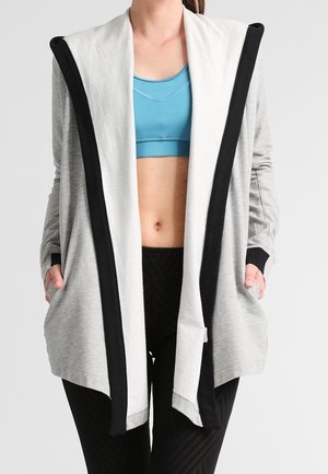 Frau, die eine hellgraue, offene Strickjacke mit schwarzem Rand über einem blauen Sport-BH und schwarzen Leggings trägt, die Hände in den Taschen.