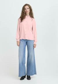 Vrouw met lang golvend haar, gekleed in een lichtroze blouse met knopen en een hoge taille met wijde lichtblauwe jeans en zwarte enkellaarsjes.