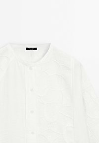 Camicia bianca con colletto rotondo caratterizzata da intricati motivi circolari e geometrici testurizzati sulla parte frontale e sulle maniche.