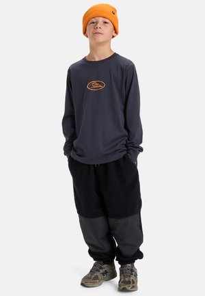 Junge mit orangefarbener Mütze, dunklem Langarmshirt mit Logo, schwarzen Fleece-Hosen und braunen New Balance Turnschuhen, steht mit den Händen in den Taschen.