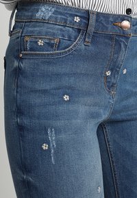 Spijkerbroek in medium blauw met versleten details en witte bloemenborduursels verspreid over de stof, met een standaard vijf-pocket ontwerp.