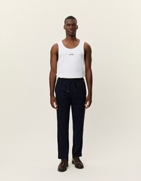 Biely tielko s logom, námornícke nohavice s šnúrkou a tmavé topánky. Model stojí vzpriamene na neutrálnom pozadí.