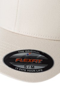 Beżowa czapka Flexfit z teksturowanej tkaniny. Zawiera czarny i pomarańczowy znaczek z logo, na którym widnieje napis "THE ORIGINAL FLEXFIT S/M IT FITS YOUR LIFE."
