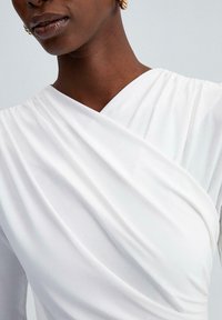 Blouse blanche drapée avec un col en V et un tissu doux et fluide. Présente un léger froncé aux épaules pour ajouter de la texture et du mouvement.