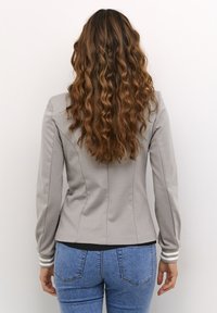 Grå tætsiddende jakke med lange ærmer, elegante linjer og ribbede manchetter. Båret over blå jeans med en glat tekstur og diskrete sømme.