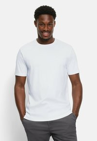 Weißes Kurzarm-T-Shirt mit Rundhalsausschnitt, aus weichem Stoff, getragen mit den Händen in den Taschen grauer Hosen. Einfaches, minimalistisches Design.