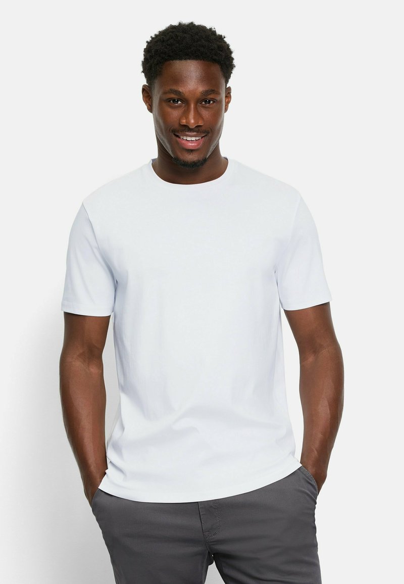 Weißes Kurzarm-T-Shirt mit Rundhalsausschnitt, aus weichem Stoff, getragen mit den Händen in den Taschen grauer Hosen. Einfaches, minimalistisches Design.