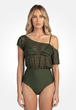 Kobieta w oliwkowozielonym jednoczęściowym kostiumie kąpielowym z przezroczystym górnym fragmentem we wzór zygzaka, z odkrytym ramieniem i cienkim paskiem po jednej stronie na białym tle.