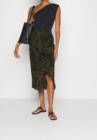 Jednokomorový černý top spárovaný s sukní délky ke kolenům v olivově zelené barvě s volány a bočním rozparkem. Černá kabelka a ploché sandály.