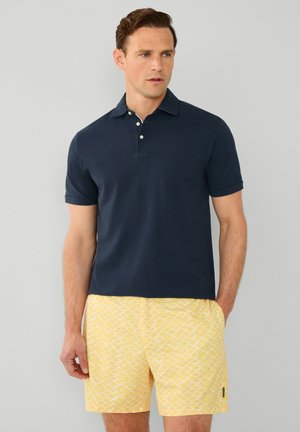 Marineblaues Kurzarm-Poloshirt mit drei Knöpfen, kombiniert mit gelben Shorts mit einem weißen Fischschuppenmuster. Schlichtes und lässiges Design.