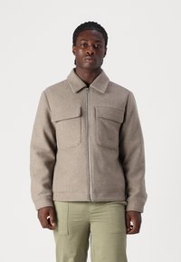 Bruine wolmix jas met een kraag, voorrits en twee borstzakken. Heeft een aansluitend design en een soepele textuur. Gepaard met groene broek.