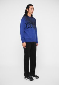 Kék kötött pulóver fekete "VERSACE JEAN COUTURE" felirattal, texturált felület; fekete nadrággal és fekete tornacipővel párosítva.