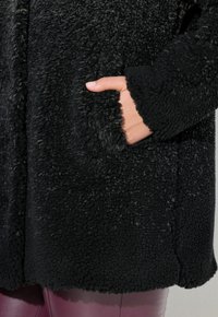 Mustan fleece-takin teksturoidulla pinnalla, jossa on sivutasku ja pyöristetty helma. Hihojen resorit ovat pehmeät ja hieman rispaantuneet.