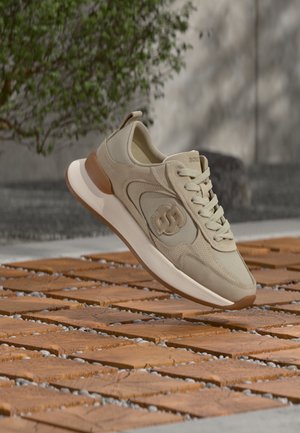Beiger und brauner Sneaker mit Logo an der Seite, Schnürsenkel gebunden, über strukturiertem Holz- und Steinboden im Freien präsentiert.