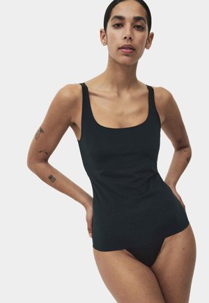 Frau mit kurzen dunklen Haaren, die ein schwarzes Tanktop und schwarze Unterwäsche trägt, steht mit den Händen in der Hüfte vor einem schlichten weißen Hintergrund.