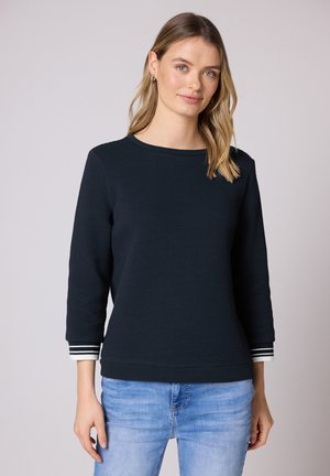 Femme aux cheveux blonds portant un haut noir texturé à manches longues avec des poignets rayés et un jean bleu clair, debout devant un fond uni.