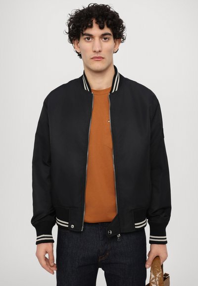 Giubbotto bomber nero con colletto e polsini a righe bianche, chiusura con zip, indossato sopra una t-shirt marrone, abbinato a jeans scuri.