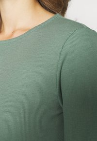 Top verde a maniche lunghe con scollatura rotonda. Il tessuto presenta un aspetto testurizzato e una silhouette aderente.