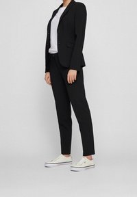 Svart skräddarsydd kavaj med två knappar, matchande svarta smala byxor och vita kanvas sneakers med subtila blå detaljer nära sulan.