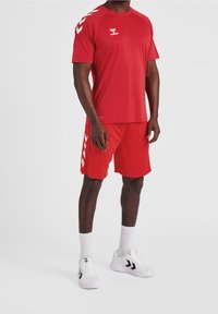 Conjunto de camiseta atlética roja y pantalones cortos, con acentos en blanco contrastantes en los hombros y franjas laterales. El material parece ligero y suave.