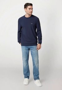 Marineblauer Pullover mit langen Ärmeln und Rundhalsausschnitt; mit kleinem Logo auf der Brust. Kombiniert mit hellblauen Jeans und weißen Turnschuhen.
