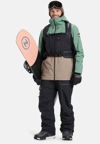 Snowboarder wearing a two-tone jacket (green and black) over brown pants. Holds a pink snowboard with a blue logo. Wears black gloves and goggles.