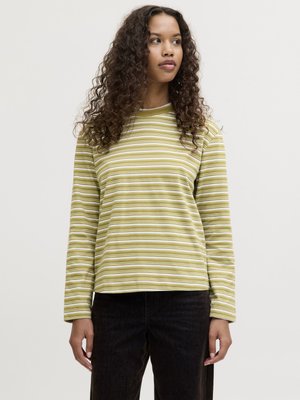 Mujer con cabello rizado que lleva una camisa de rayas largas verde y blanca y pantalones negros, de pie contra un fondo liso y claro.
