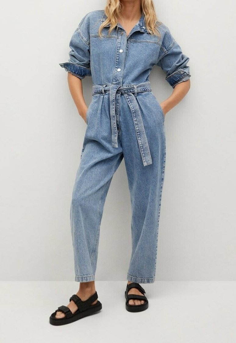 Personne portant une combinaison en denim bleu clair avec les manches retroussées et une ceinture assortie, associée à des sandales noires à bout ouvert, debout contre un mur uni.