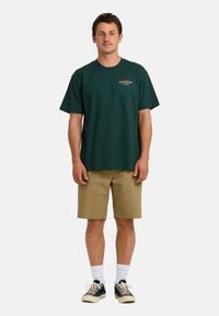 Camiseta de algodón verde oscuro de mangas cortas, combinada con pantalones cortos beige. La camiseta presenta un pequeño logo horizontal en el pecho.