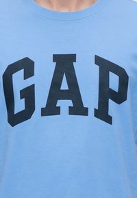 Светлосиня риза с големи черни главни букви, изписващи GAP през гърдите.