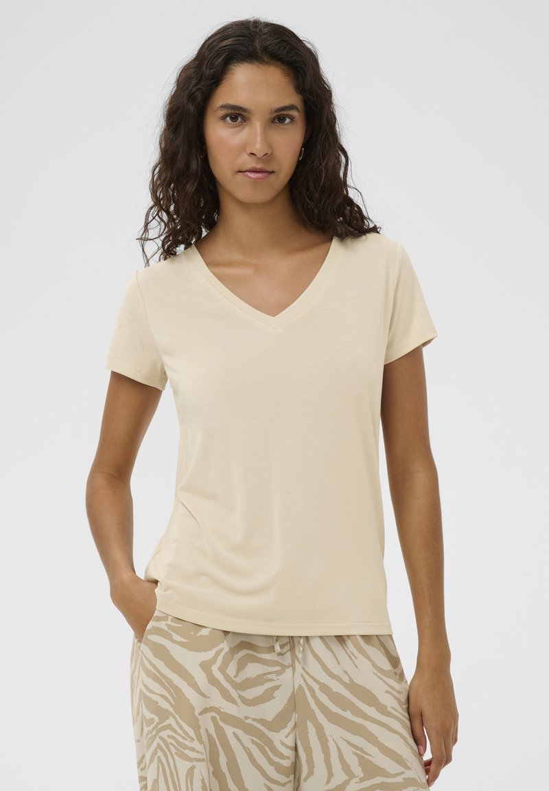 Camiseta beige de manga corta con cuello en V, confeccionada con tela suave y lisa. Presenta un ajuste relajado con un brillo sutil. Combinada con pantalones beige estampados.
