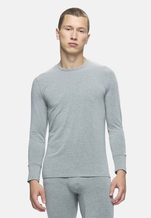 Jeune homme portant un haut à manches longues gris clair et un pantalon ajusté assorti sur fond blanc uni.