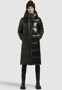 Svart, blank, vadderad lång jacka med hög krage, utrustad med sidofickor och dragkedja fram. Buren över svarta leggings och stövlar.