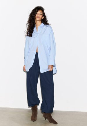 Femme aux cheveux longs et bouclés portant une chemise oversize bleu clair, un pantalon ample bleu foncé et des bottines marron à talons, debout contre un mur blanc.