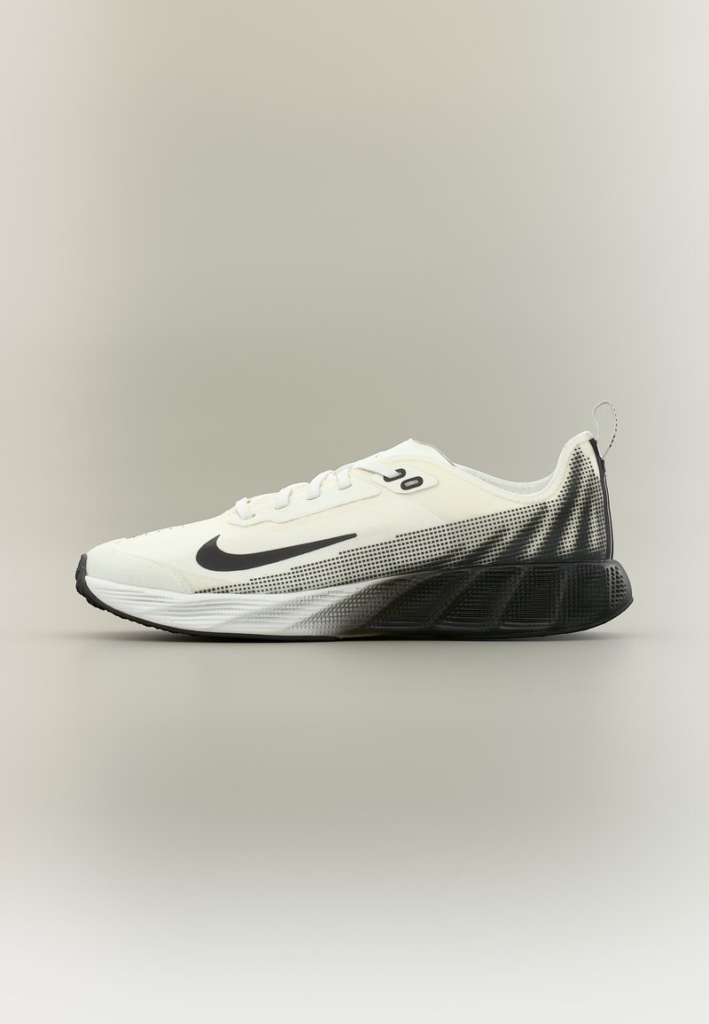 Λευκό και μαύρο αθλητικό παπούτσι Nike με διαβαθμισμένο πουά σχέδιο στη φτέρνα και μαύρη σόλα, εμφανίζεται από το πλάι σε ουδέτερο φόντο.