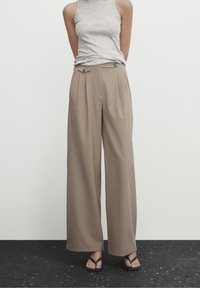 Pantalon large en tissu beige clair, doté d'une taille haute avec plis et double bouton. Associé à un haut sans manches gris et des sandales foncées.